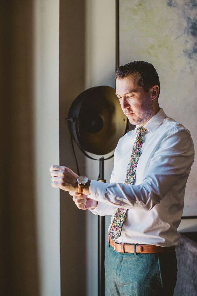 groom in white shirt with bright floral tie puts watch on while getting ready for wedding day