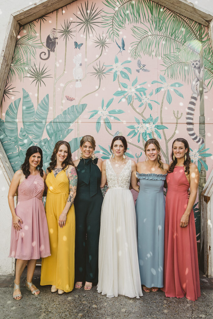 bride in embellished deep v neck wedding dress with a chiffon skirt with bridesmaids in rainbow colored mixed dresses stand in front of pink key hole wall at Valentine DTLA