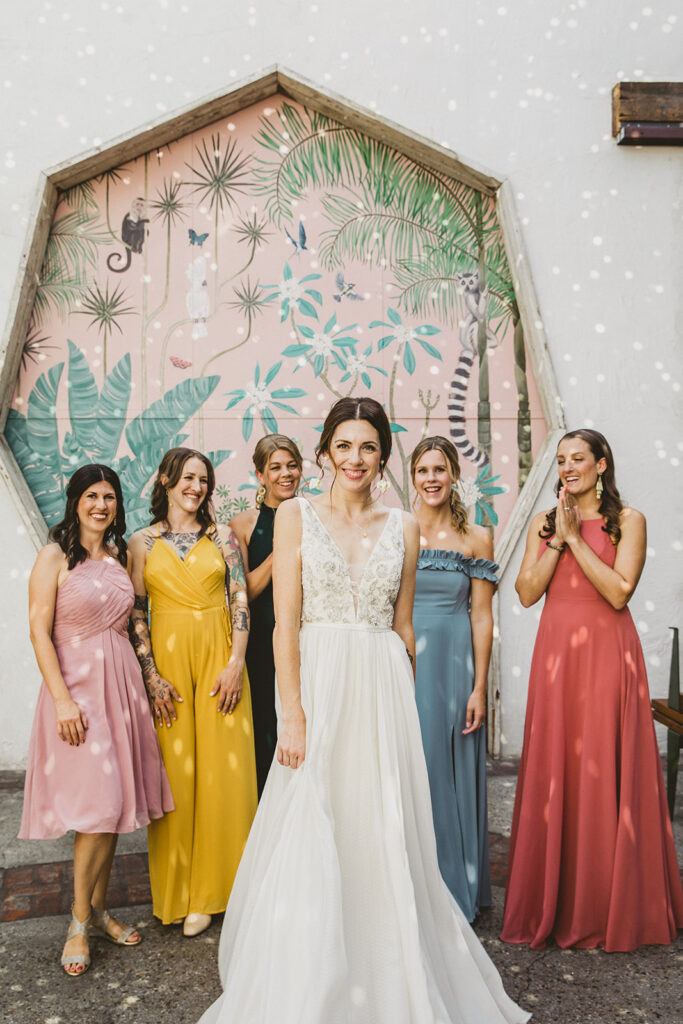 bride in embellished deep v neck wedding dress with a chiffon skirt with bridesmaids in rainbow colored mixed dresses stand in front of pink key hole wall at Valentine DTLA