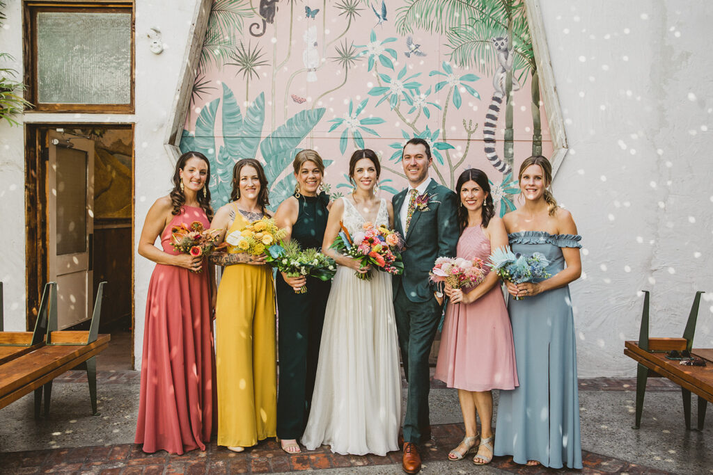 bride in embellished deep v neck wedding dress with a chiffon skirt and groom in custom green suit with bright floral tie stands with wedding party in front of pink keyhole wall at Valentine DTLA