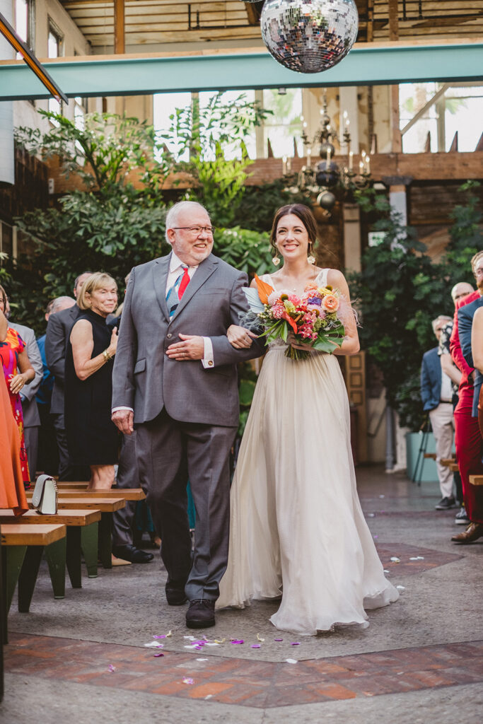 bride in embellished deep v-neck wedding dress with chiffon skirt holding bright jungle themed floral bridal bouquet walks down ceremony aisle at Valentine DTLA