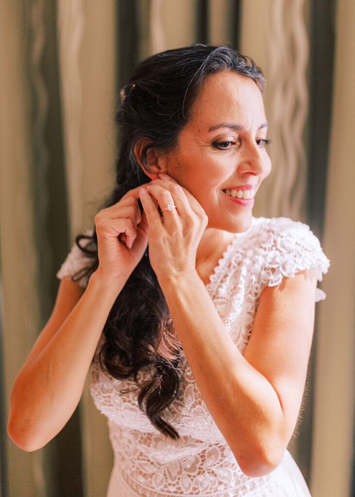 bride in lace bodice wedding dress putting on earrings 