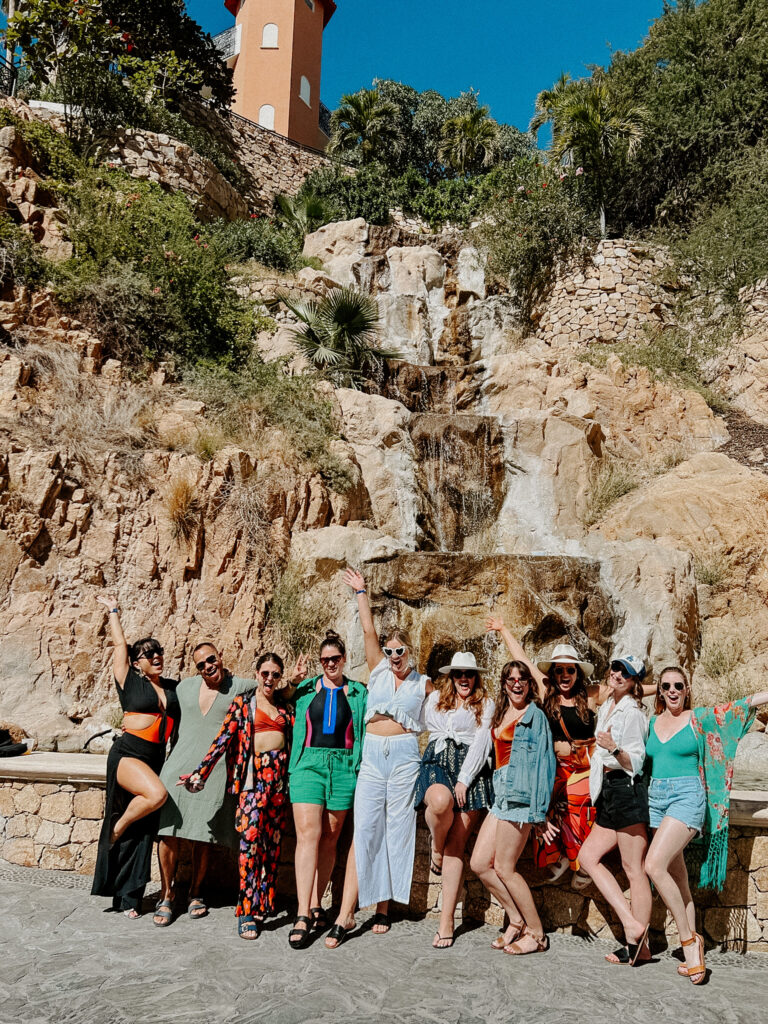 bride to be in white matching linen set and heart shaped sunglasses stands with friends in front of waterfall in Cabo