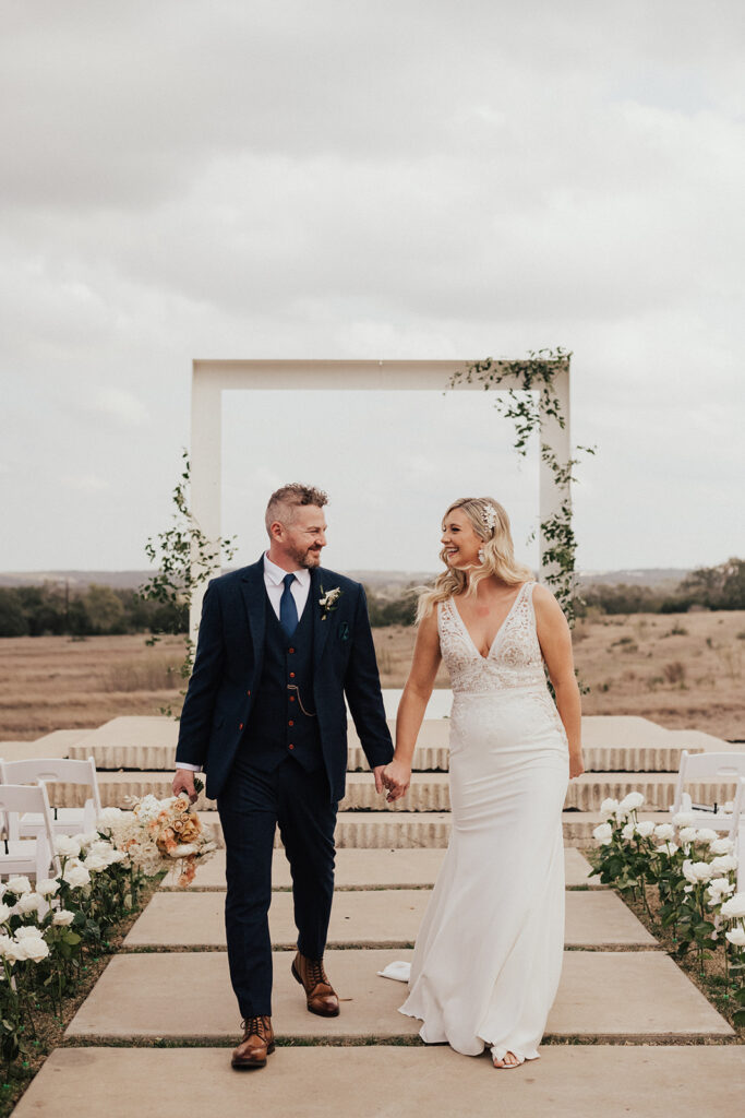 bride in deep v neck wedding dress with lace cut out and groom in navy blue suit in front of framed wedding ceremony arch with greenery before destination wedding ceremony