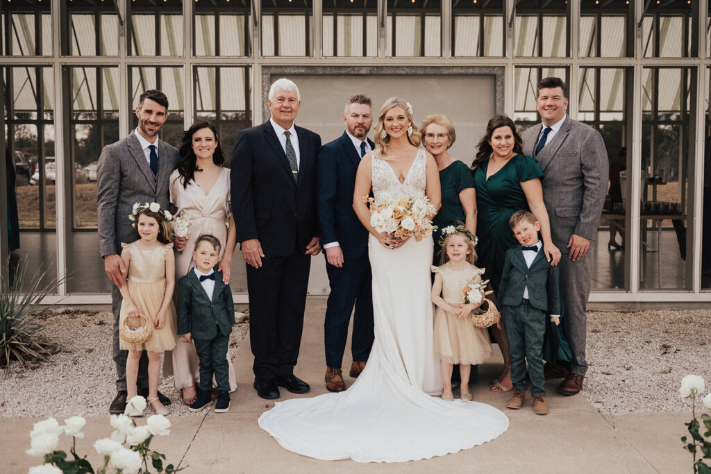bride in deep v neck lace wedding dress and groom in navy blue suit stand with family in green, champagne and charcoal grey attire 