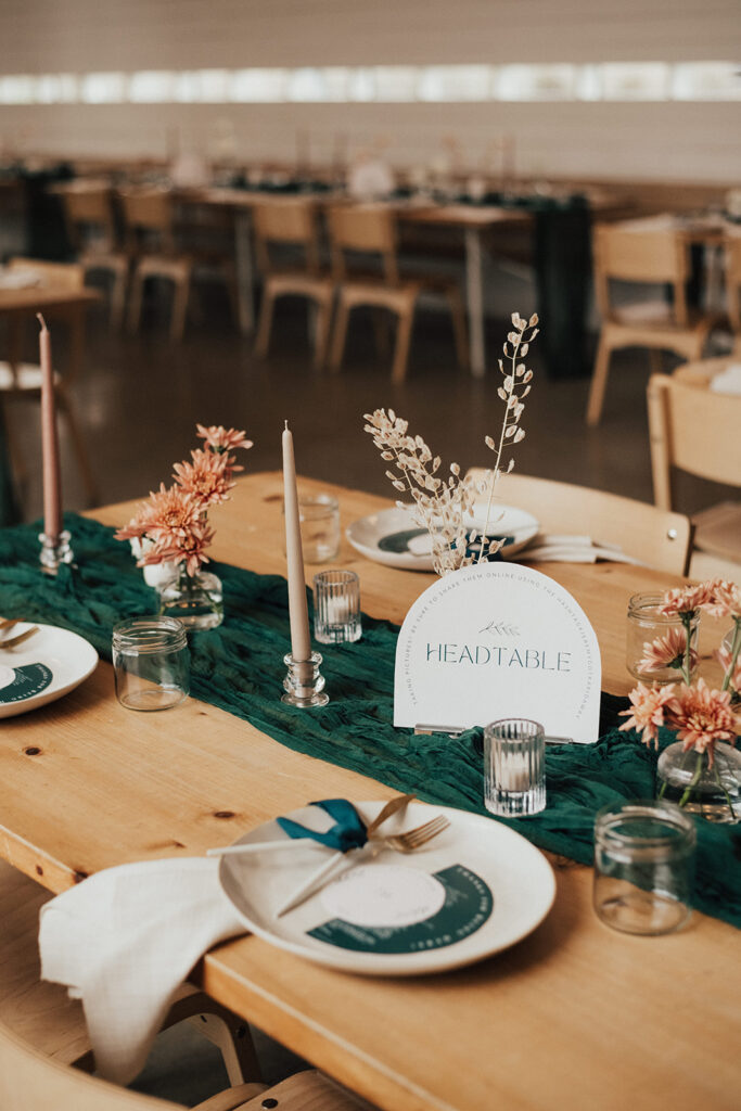Modern wedding reception at Prospect House with wooden oak chairs and tables, dark green linens, beige taper candles and small bud vases. Rounded head table sign