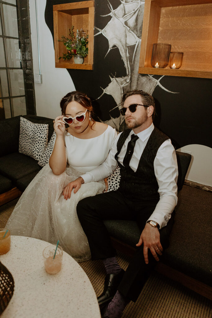 bride in two piece wedding reception outfit and groom in black suit cheers while wearing sunglasses