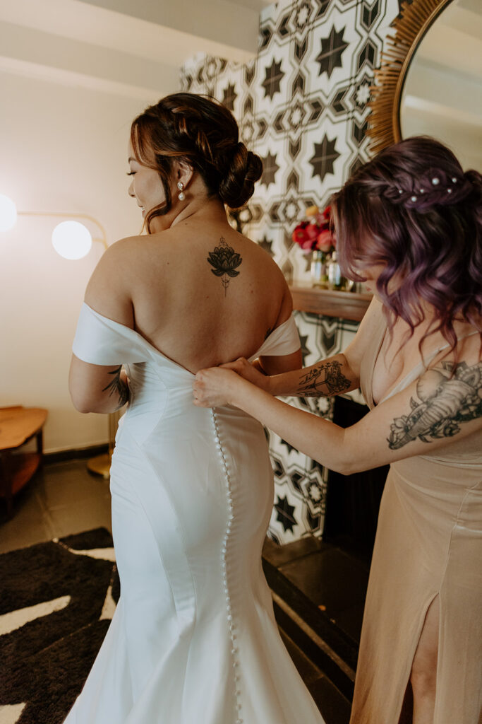 bride with lotus flower tattoo gets into minimalist off shoulder wedding dress with buttons