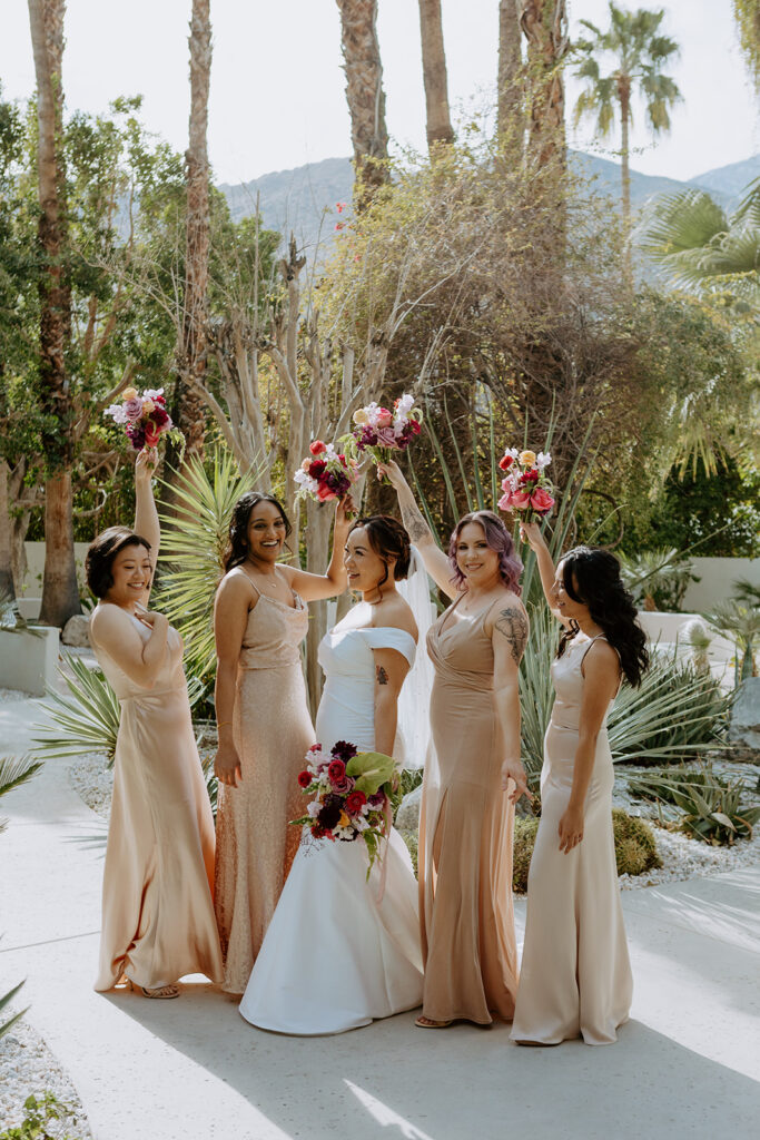 bride in minimalist off shoulder wedding dress holds colorful bouquet with bridesmaids in beige satin bridesmaid dresses
