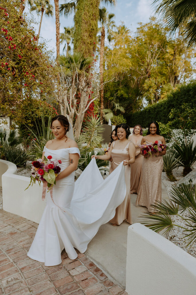 bride in minimalist off shoulder wedding dress walks with bridesmaids holding her train 