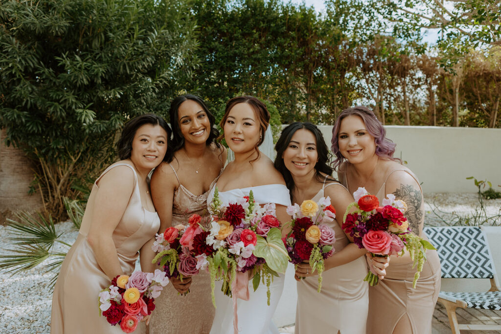 bride in minimalist off shoulder wedding dress holds colorful bouquet with bridesmaids in beige satin bridesmaid dresses