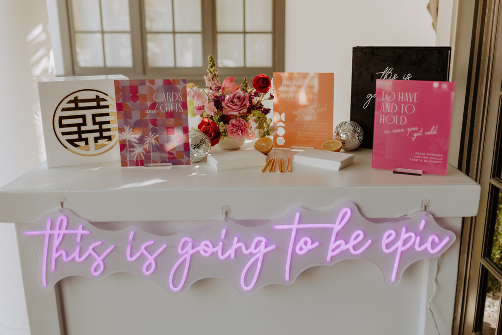 pool side wedding welcome table with custom neon sign that reads "This  is going to be epic" with colorful mid-century inspired signs and baskets with blankets