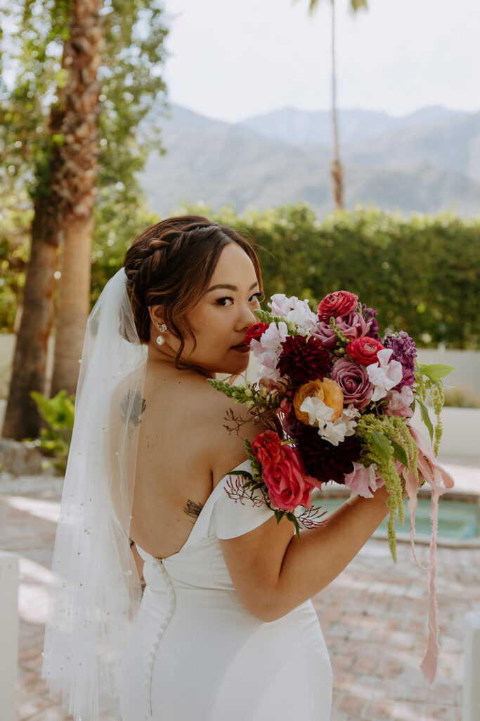 bride in minimalist off shoulder wedding dress with colorful bouquet and veil with pearls poses for portrait shots in Palm Springs