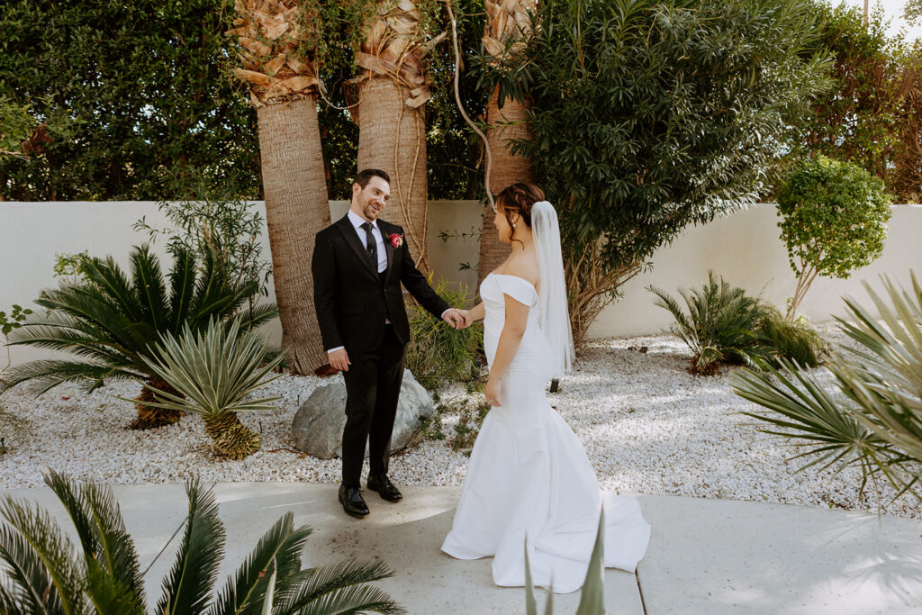 bride in minimalist off shoulder wedding dress with veil with pearls has first look with groom in black suit in Palm Springs