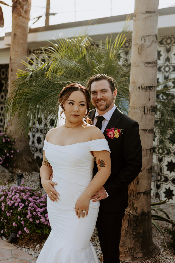 bride in minimalist off shoulder wedding dress with veil with pearls has first look with groom in black suit in Palm Springs