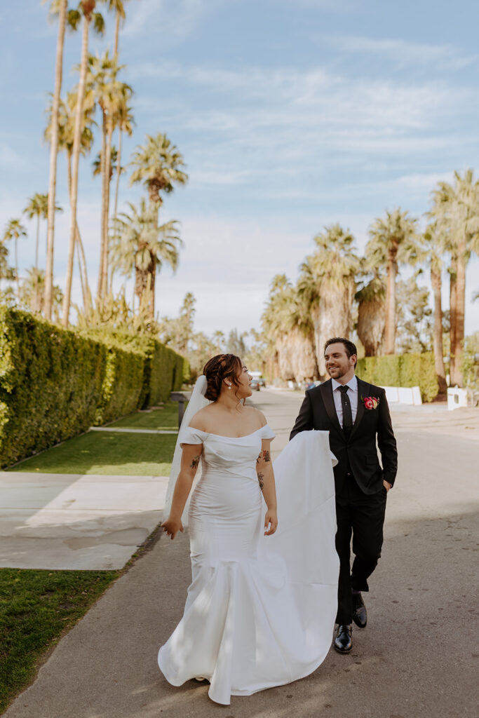 bride in minimalist off shoulder wedding dress with veil with pearls walks with groom in black suit in Palm Springs