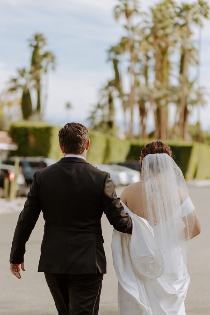 bride in minimalist off shoulder wedding dress with veil with pearls walks with groom in black suit in Palm Springs
