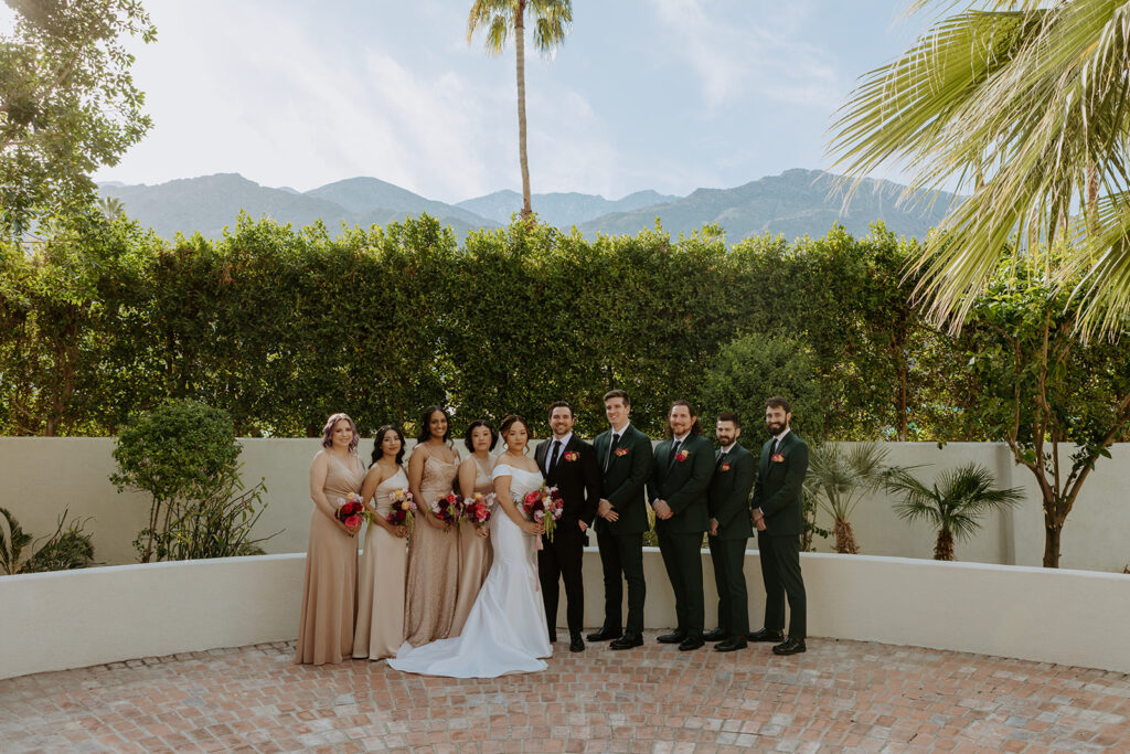 bride in minimalist off shoulder wedding dress with pearl embellished veil and bridesmaids in beige dresses stands with groom in black suit and groomsmen in dark green suits in Palm Springs