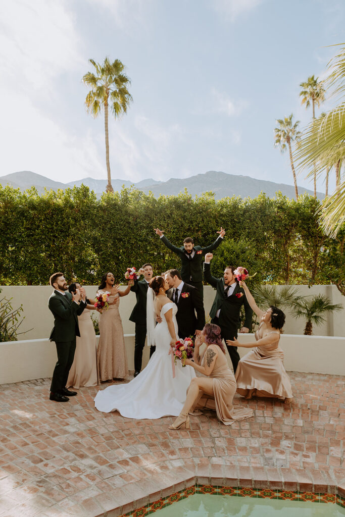 bride in minimalist off shoulder wedding dress with pearl embellished veil and bridesmaids in beige dresses stands with groom in black suit and groomsmen in dark green suits in Palm Springs