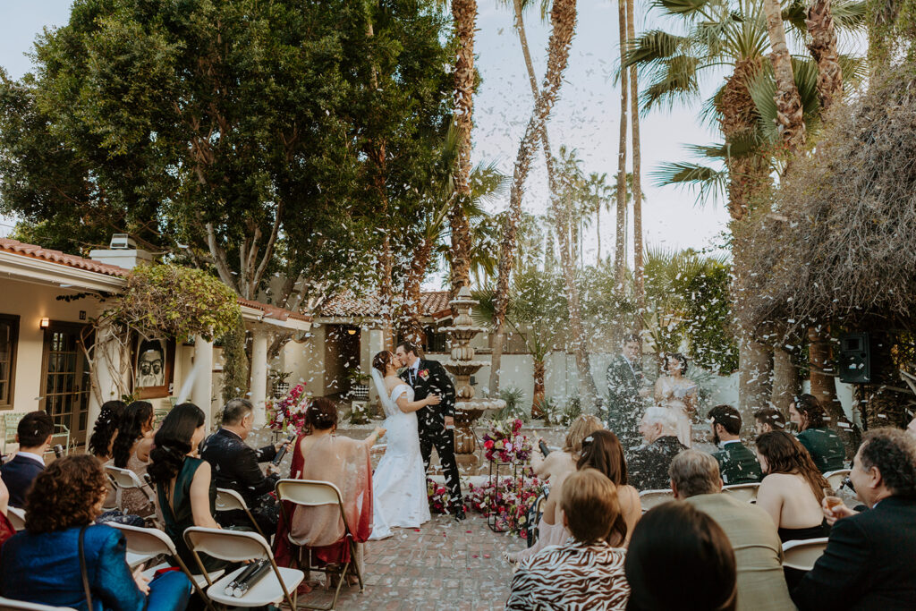 bride and groom first kiss during colorful wedding ceremony in front of fountain at Villa Royal Palm Springs while confetti is popped by guests