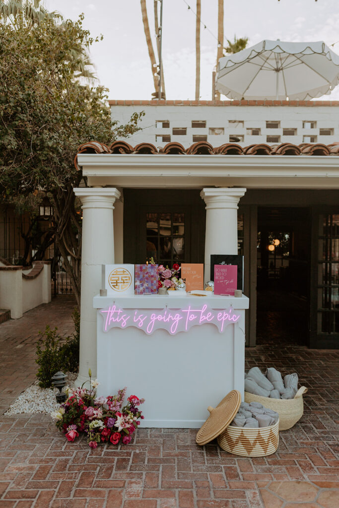 pool side wedding welcome table with custom neon sign that reads "This  is going to be epic" with colorful mid-century inspired signs and baskets with blankets