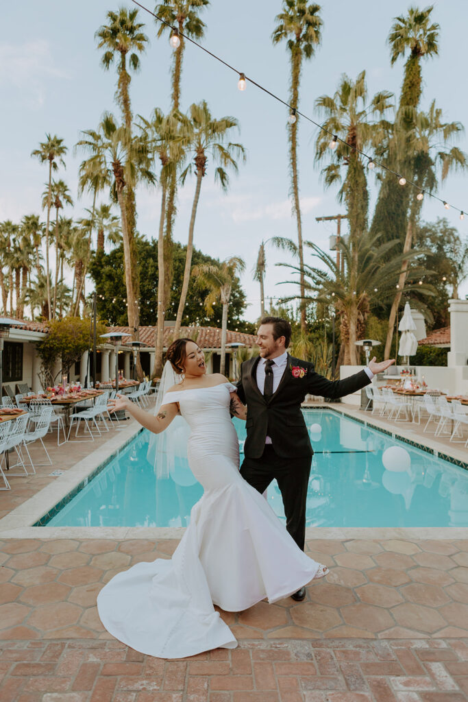 bride in minimalist off shoulder wedding dress and groom in black suit stand in front of pool side wedding reception at Villa Royale Palm Springs