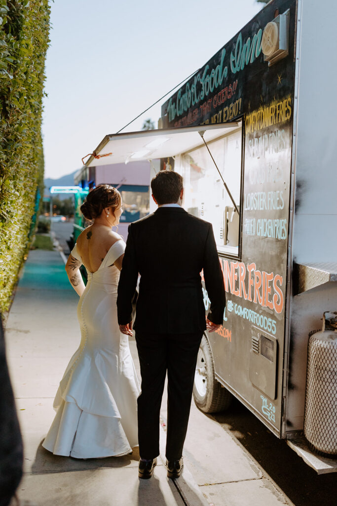 bride in minimalist off shoulder wedding dress and groom in black suit get food at food truck