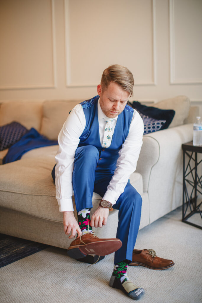 groom in blue suit and cartoon socks putting on brown shoes 