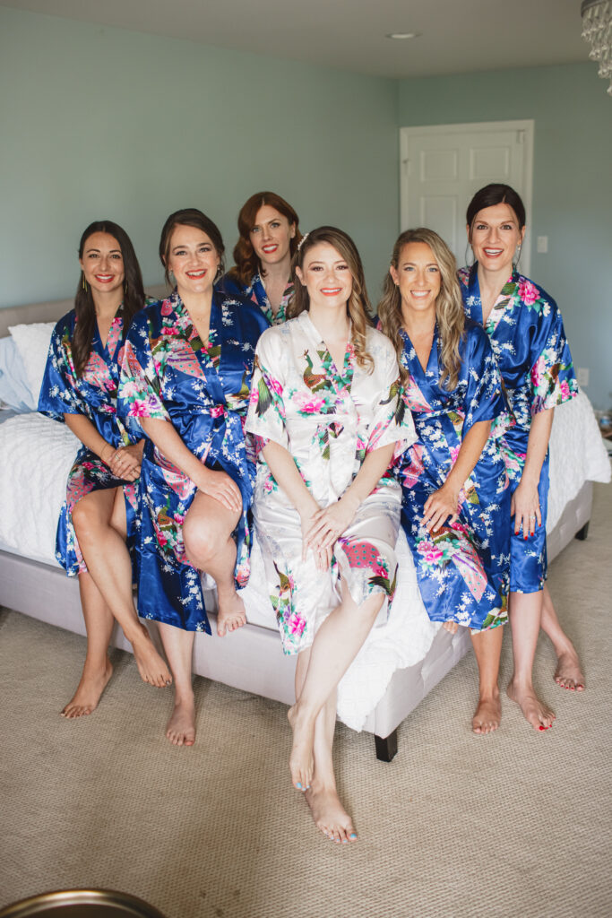 bride in white floral robe sits on bed with bridesmaids in blue floral robes