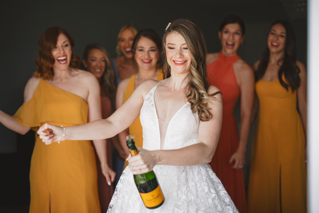 bride in deep vneck lace wedding dress with tiered skirt opens champagne with bridesmaids in sunset colored dresses