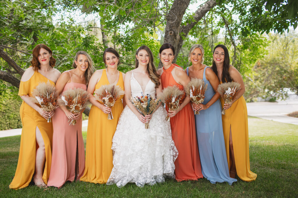 bride in lace vneck wedding dress with tiered skirt stands with bridesmaids in sunset colored dresses and dried floral bouquets