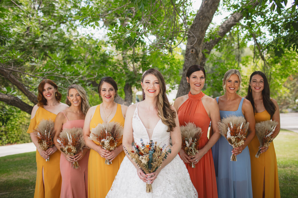 bride in lace vneck wedding dress with tiered skirt stands with bridesmaids in sunset colored dresses and dried floral bouquets