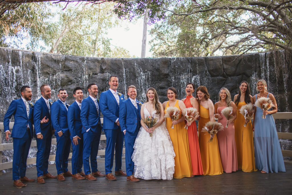 bride in lace deep vneck wedding dress with tiered skirt and groom in blue suit stands with bridal party in sunset colored attire at the Oak Room 