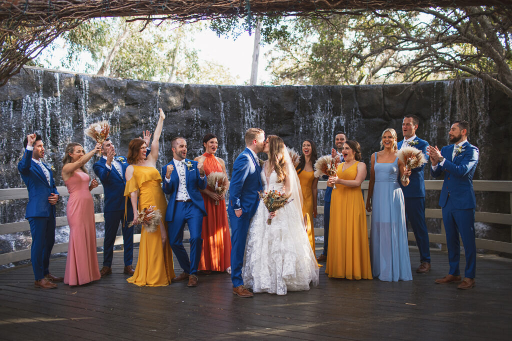 bride in lace deep vneck wedding dress with tiered skirt and groom in blue suit stands with bridal party in sunset colored attire at the Oak Room 