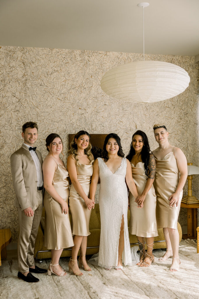 bride with lace wedding dress and leg slit stands with co-ed wedding party in Champagne colored dress and suit