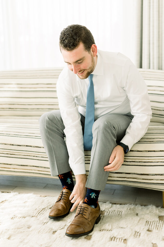 groom putting on brown shoes with white shirt, grey pants and blue tie