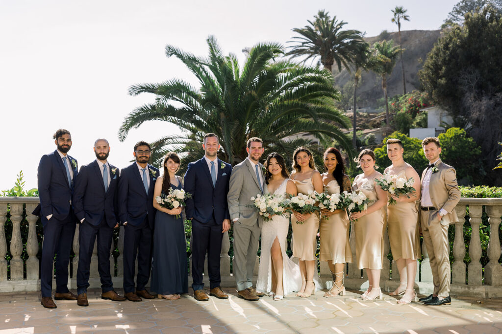 bride in lace wedding dress with leg slit stands with groom in grey suit and blue tie with their wedding party in champagne and navy blue colored suits and dresses at Bel-Air Bay Club