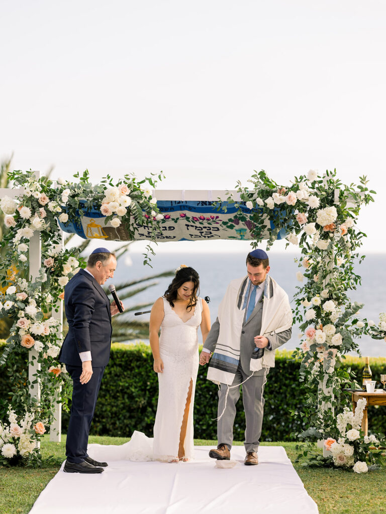 groom in light grey suit with blue tie stomps on glass with bride in lace wedding dress during Jewish wedding ceremony at Bel Air Bay Club with ocean view 