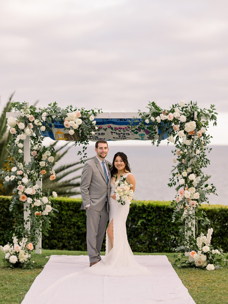bride in deep v-neck lace dress with leg opening and long train stands with groom in light grey suit and blue tie in front of their chuppah at Bel Air Bay Club