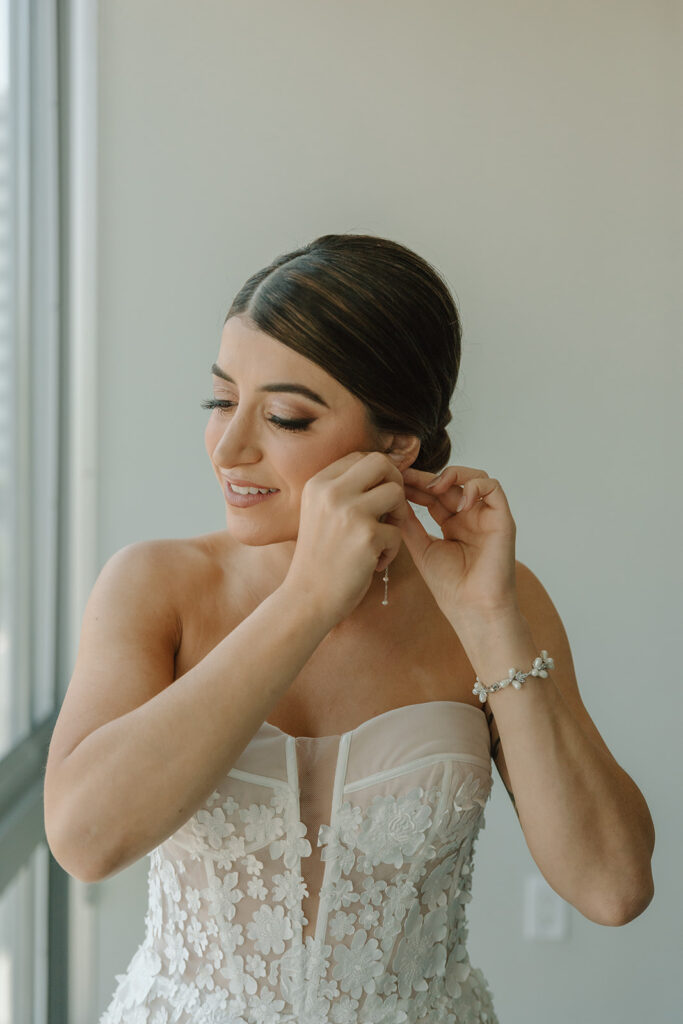 bride in low slicked back bridal bun wearing strapless wedding dress with 3d floral appliqué putting on pearl drop earrings