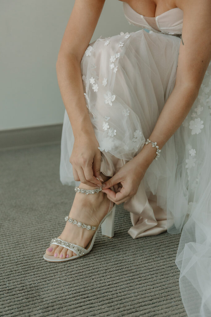 bride in low slicked back bridal bun wearing strapless wedding dress with 3d floral appliqué putting bridal shoes