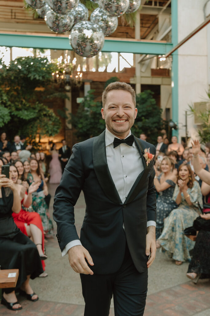 groom dances down ceremony aisle with disco ball installation behind him 