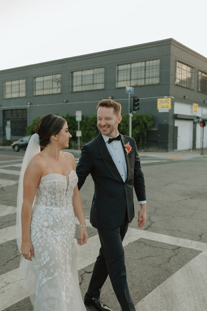 bride in strapless wedding dress with 3d floral appliqué and pink tropical bridal bouquet walks with groom in black tuxedo in DTLA