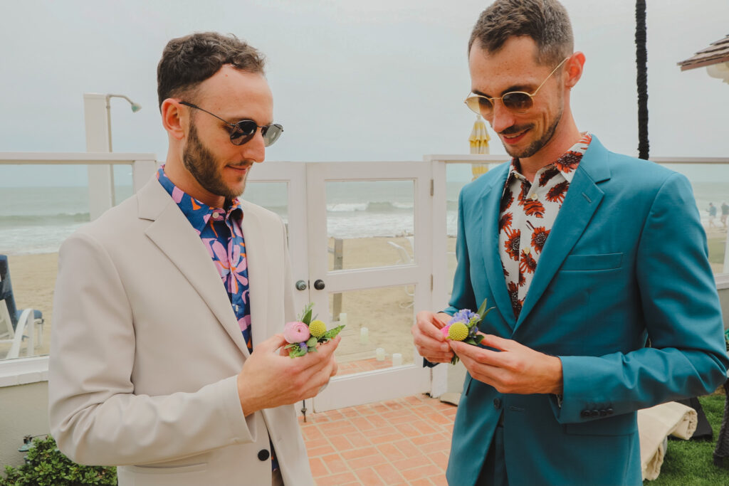 groom in bold print shirt and tan suit putting on boutonniere with groom in bold print shirt and blue suit