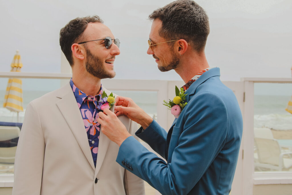 groom in bold print shirt and tan suit putting on boutonniere with groom in bold print shirt and blue suit
