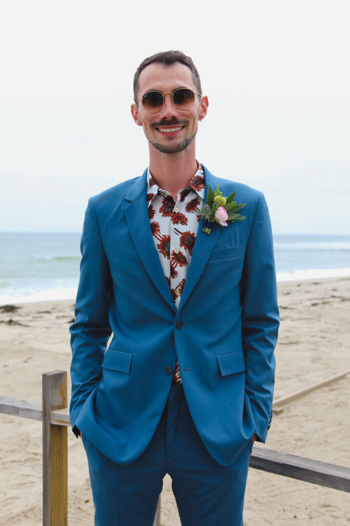 groom in bold print shirt and blue suit with colorful boutonniere stands on beach for private beach wedding ceremony