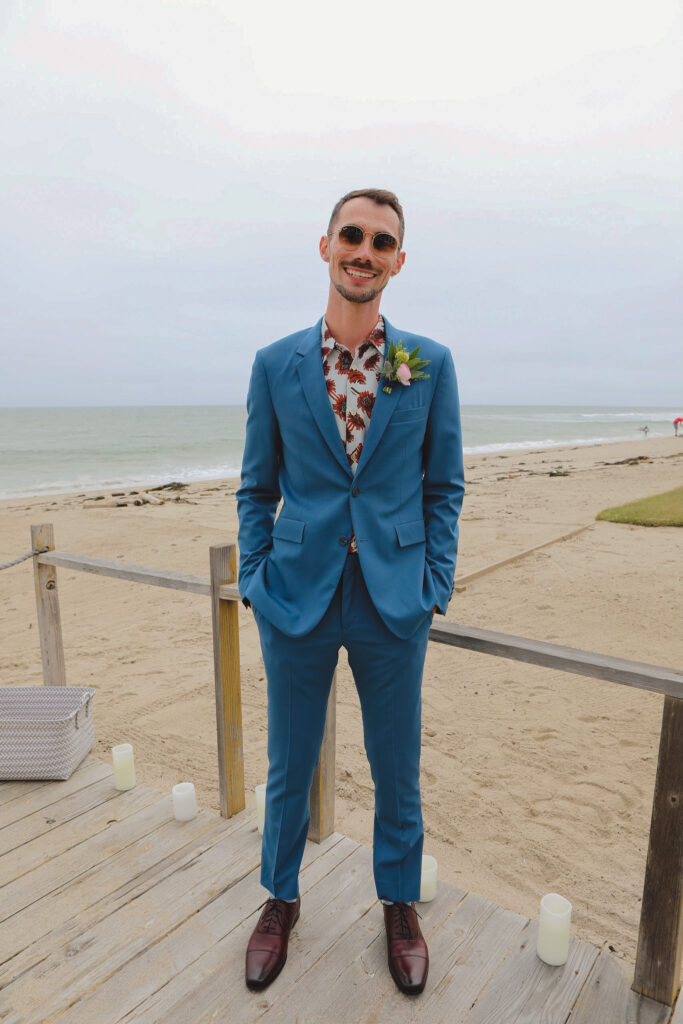 groom in bold print shirt and blue suit with colorful boutonniere stands on beach for private beach wedding ceremony