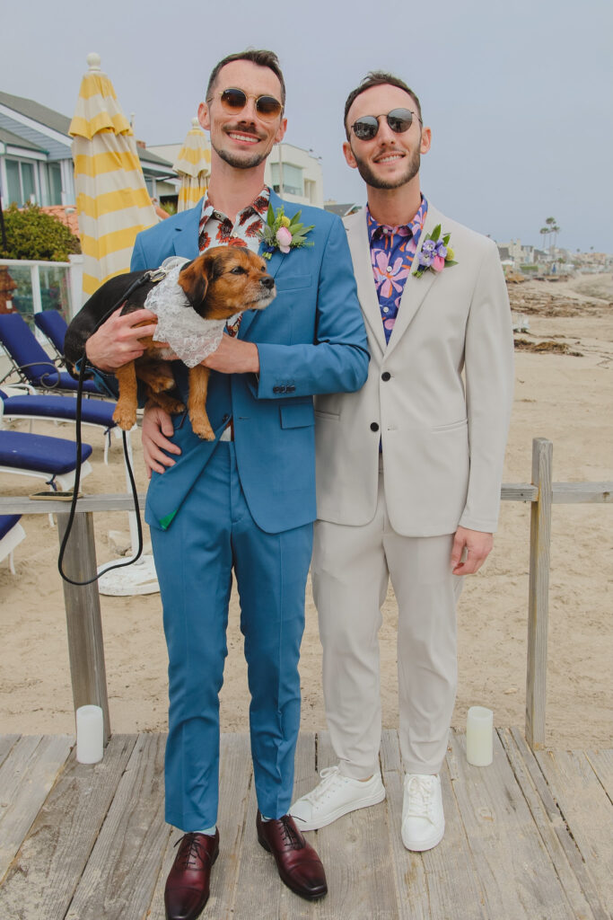 groom in bold print shirt and blue suit and groom in bold print shirt with tan suit hold dog with flower collar stand on beach for private beach wedding ceremony