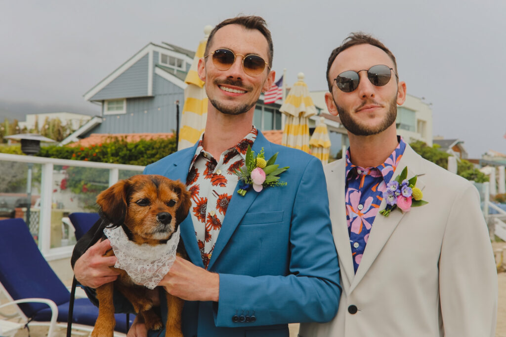 Groom in bold print shirt and tan suit with groom in bold print shirt and blue suit stands with dog in floral collar for private beach wedding