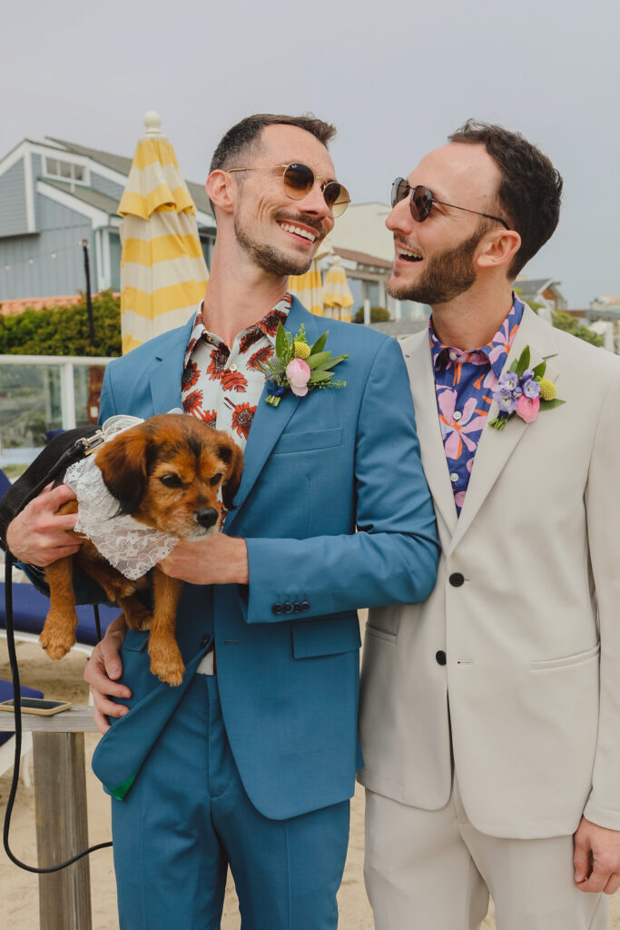 groom in bold print shirt and blue suit and groom in bold print shirt with tan suit hold dog with flower collar stand on beach for private beach wedding ceremony